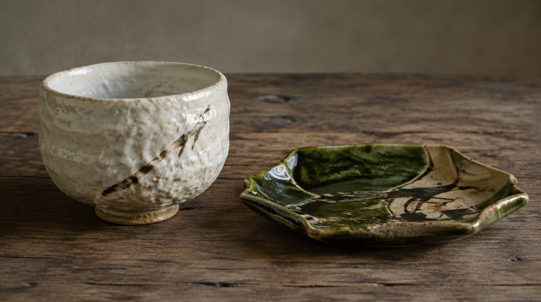 A white Shino tea bowl with an iron brushstroke beside a green-glazed Oribe dish on a wooden table