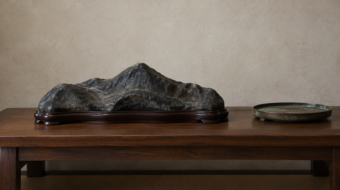 A dark natural viewing stone shaped like a distant mountain range on a carved wooden base, beside a shallow bronze tray