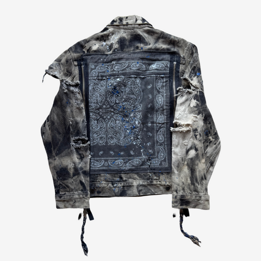 A heavily distressed denim jacket shown from the back, featuring a large dark blue paisley bandana patch on the back panel and a mottled black-and-white bleach wash. The jacket has ripped details and drawstring ties hanging from the hem.